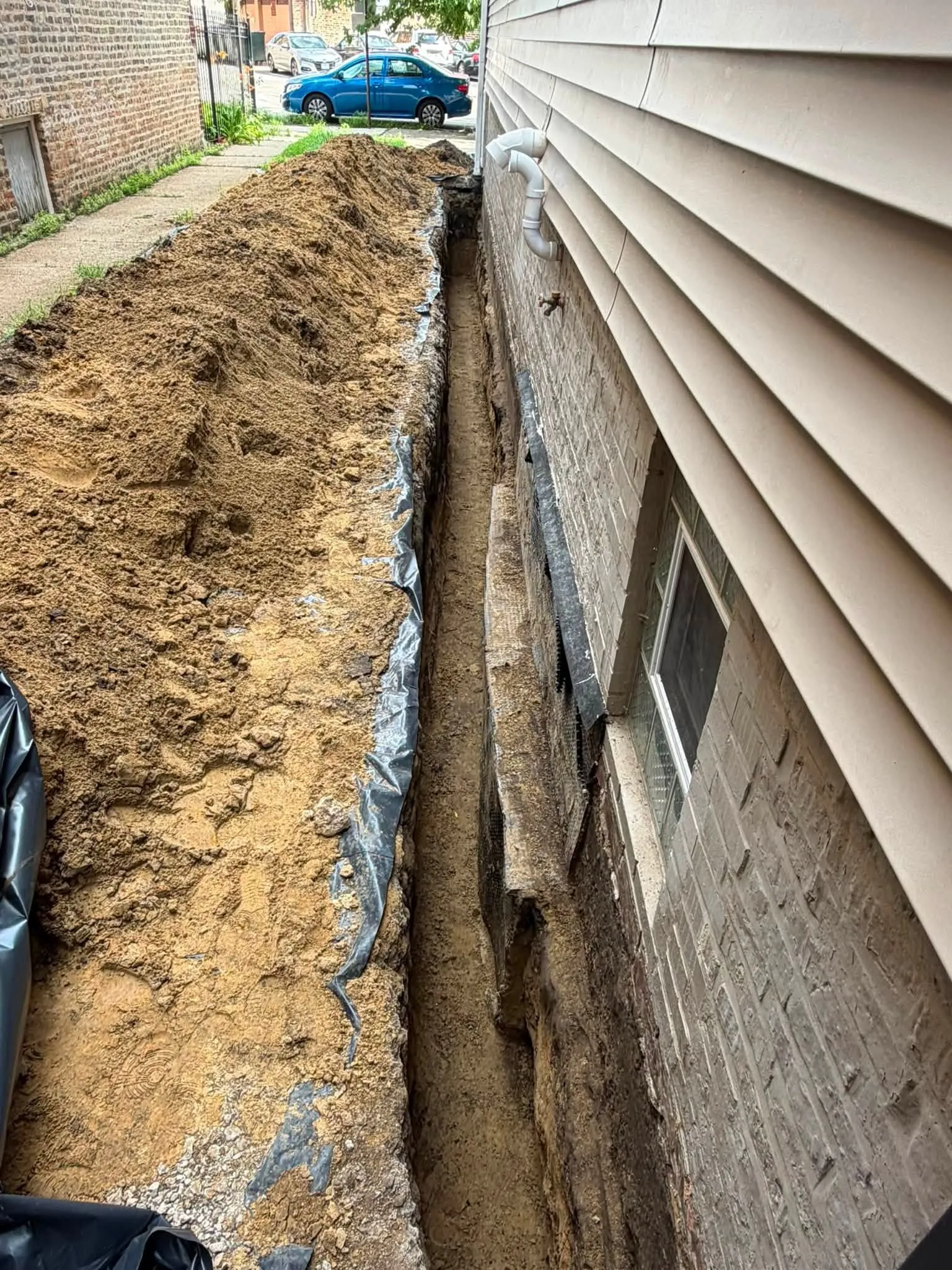 Open drainage trench alongside a house with poly sheeting and downspout connection, Toronto